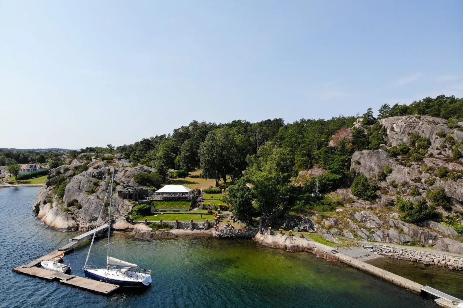 Alaskaträdgården med översiktlig by över stenträdgården med blommor och stora gräsytor och skog intill vatten gränsen och småbåtshamnen på Nord-långö.